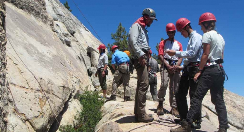 A group of people wearing helmets and other safety gear gather at the base of a rock wall, preparing to climb